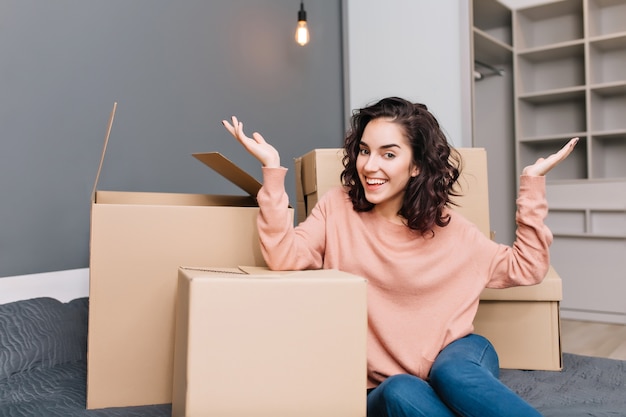 Aufgeregte junge Frau auf Bettumrandungskästen, Karton lächelnd in der modernen Wohnung. Umzug in eine neue Wohnung, Ausdruck wahrer positiver Emotionen in einem neuen Zuhause mit modernem Interieur