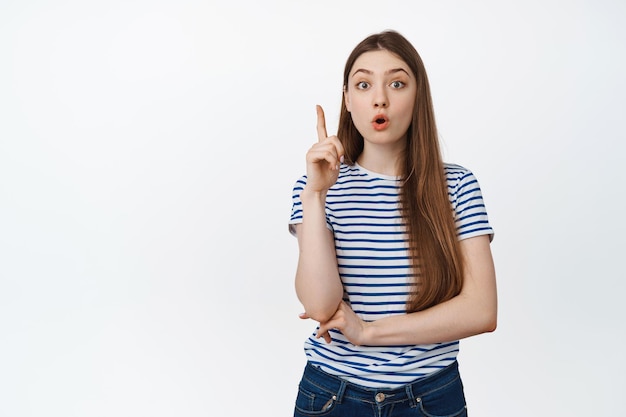 Aufgeregte junge Frau hatte eine Idee, hob die Finger-Heureka-Geste, zeigte nach oben, fand oder entdeckte etwas Interessantes, stand in gestreiftem T-Shirt auf weißem Hintergrund.