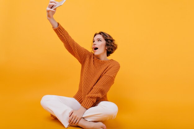 Aufgeregte junge Frau mit stilvollem Haarschnitt, der Selfie beim Sitzen mit gekreuzten Beinen macht