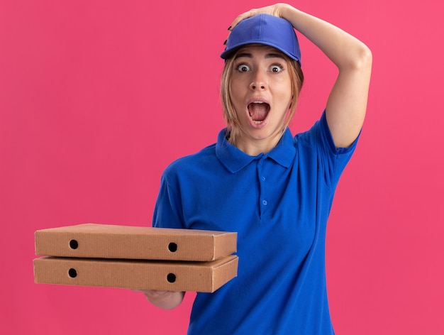 Aufgeregte junge hübsche Lieferfrau in Uniform setzt Hand auf Kappe und hält Pizzaschachteln isoliert auf rosa Wand