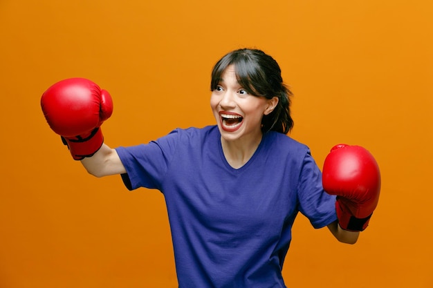 Kostenloses Foto aufgeregte junge sportliche frau mit t-shirt und boxhandschuhen, die die fäuste isoliert auf orangefarbenem hintergrund in der luft hält