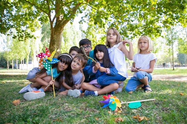 Aufgeregte Kinder, die zusammen auf Gras im Park sitzen, wegschauen, Windrad halten, Leistung beobachten. Kinderparty oder Unterhaltungskonzept