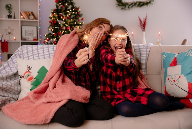 Aufgeregte Mutter und Tochter halten Wunderkerzen bedeckt mit Decke sitzen auf der Couch und genießen die Weihnachtszeit zu Hause