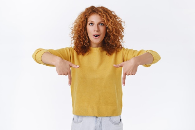 Kostenloses Foto aufgeregte, neugierige rothaarige frau mit lockigem haar trägt einen gelben pullover, der nach unten zeigt und erstaunt aussieht, angenehm überrascht und erstaunt, ein neues produkt auszuprobieren, das freunden zeigt, wo sie einen guten verkauf finden