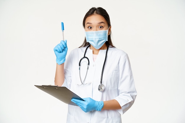 Aufgeregte Ärztin, asiatische Ärztin, die Zwischenablage hält und Stift hochhebt, Lösung oder Idee gefunden, in medizinischer Gesichtsmaske auf weißem Hintergrund stehend