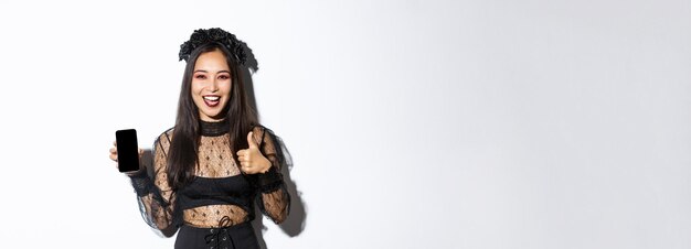 Aufgeregte und zufriedene asiatische Frau im Halloween-Kostüm, die Daumen hoch zur Zustimmung zeigt und demonstriert