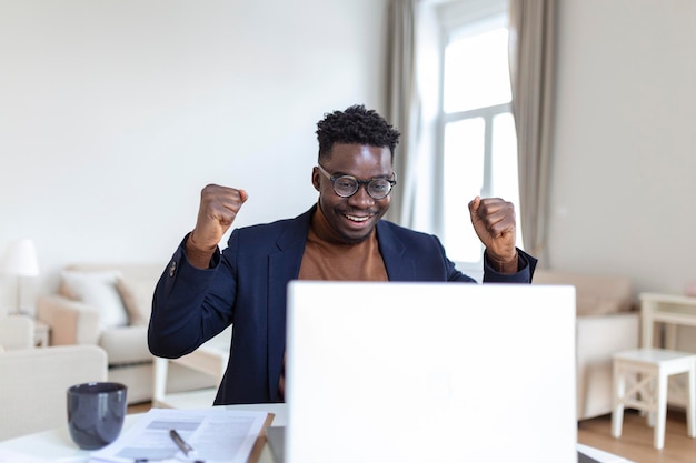 Aufgeregter afroamerikanischer Mann, der gute Nachrichten in E-Mails liest, erhält eine neue Jobbeförderung mit einem Laptop, der auf den Bildschirm schaut und vor Freude schreit und eine Ja-Geste zum Feiern zeigt