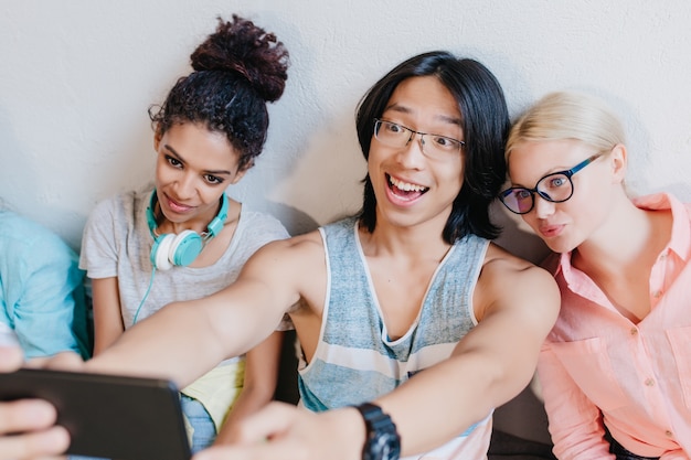 Aufgeregter asiatischer Student in der Brille, die Foto von sich macht, der zwischen blonder Frau und afrikanischer junger Frau sitzt. Stilvolle Freunde machen Selfie nach dem Test in der Universität.