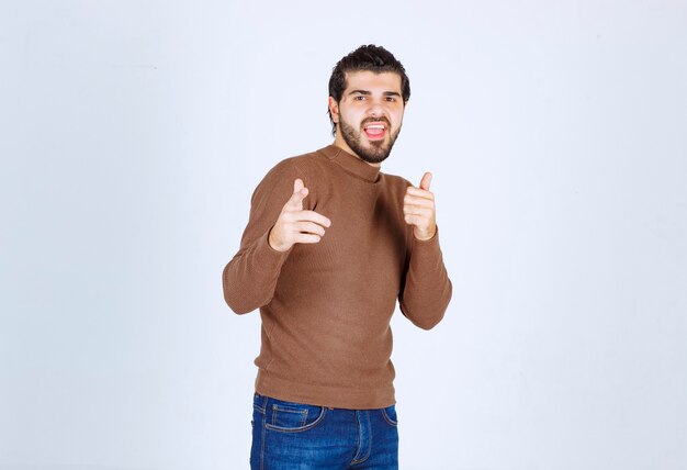 Aufgeregter bärtiger Mann im braunen Pullover, der mit den Fingern auf die Kamera zeigt, die auf einem weißen Hintergrund isoliert ist. Foto in hoher Qualität