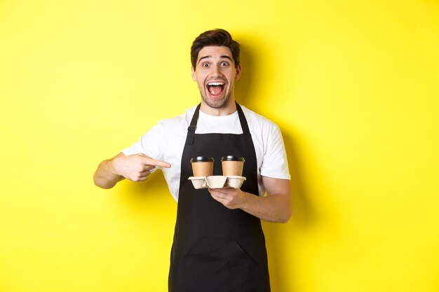 Aufgeregter Barista in schwarzer Schürze, der mit den Fingern auf Kaffeetassen zum Mitnehmen zeigt und vor gelbem Hintergrund glücklich steht.