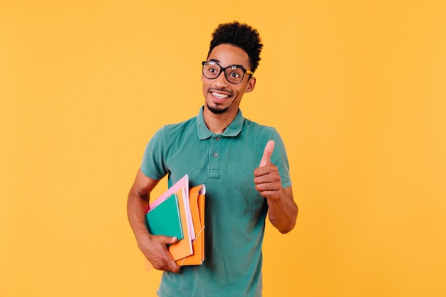 Aufgeregter braunhaariger Kerl in Gläsern, die Bücher halten. sorgloser afrikanischer Student isoliert.