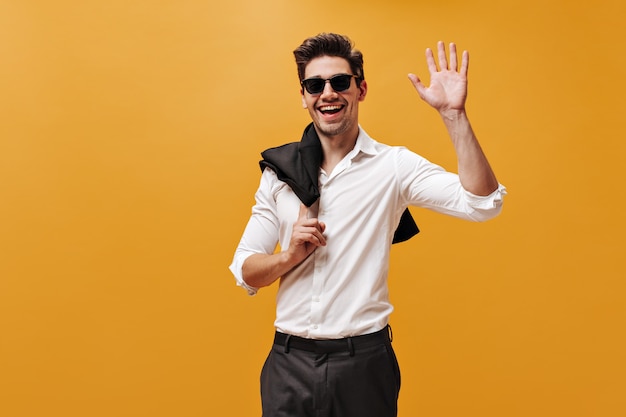 Aufgeregter charmanter Brunet-Mann in weißem Hemd, Sonnenbrille und schwarzer Hose lächelt, hält Jacke und winkt zur Begrüßung an orangefarbener Wand.