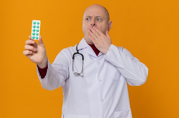 Aufgeregter erwachsener Mann in Arztuniform mit Stethoskop, das die Blisterpackung hält und betrachtet, die die Hand auf den Mund legt