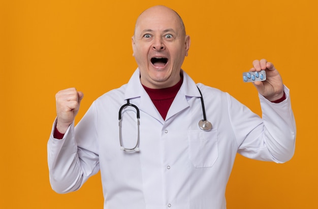 Aufgeregter erwachsener slawischer Mann in Arztuniform mit Stethoskop, der Medizinblisterpackung hält und Faust hält