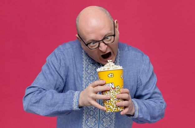 Aufgeregter erwachsener slawischer mann in blauem hemd mit optischer brille, der popcorn-eimer hält und betrachtet Kostenlose Fotos