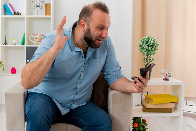 Aufgeregter erwachsener slawischer Mann sitzt auf Sessel, der Hand hebt und Telefon im Wohnzimmer betrachtet