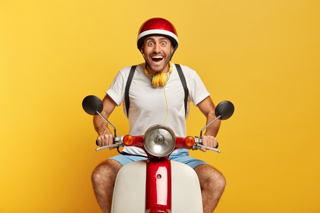 Aufgeregter glücklicher hübscher männlicher Fahrer auf Roller mit rotem Helm