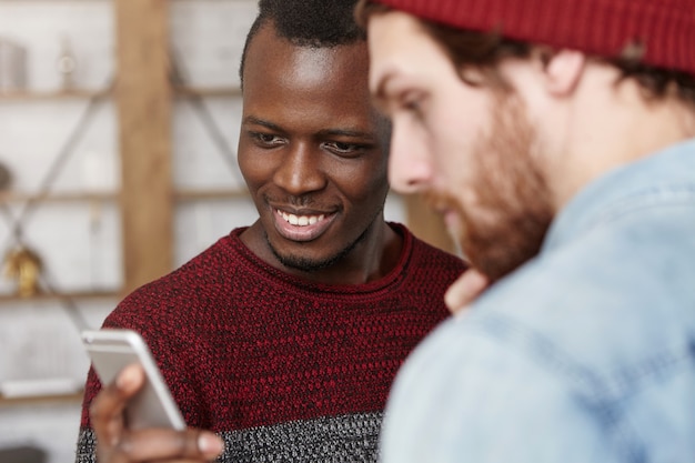 Aufgeregter glücklicher junger schwarzer Mann im Pullover, der seinen modischen weißen Freund zeigt, der Blog-Konto in sozialen Netzwerken inspiriert. Zwei beste männliche Freunde, die Handy zusammen im Café benutzen. Selektiver Fokus