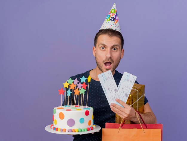 Aufgeregter gutaussehender Mann, der Geburtstagskappe hält, hält Geburtstagskuchenpapier-Einkaufstasche-Geschenkbox und Flugtickets lokalisiert auf lila Wand