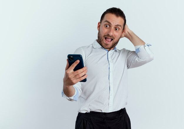 Aufgeregter gutaussehender Mann legt Hand auf Kopf hinter Blick auf Telefon lokalisiert auf weißer Wand