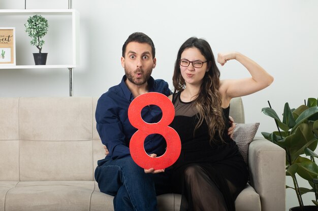 Aufgeregter gutaussehender Mann mit roter Acht und hübsche junge Frau in optischer Brille, die ihren Bizeps anspannt, der am internationalen Frauentag im März auf der Couch im Wohnzimmer sitzt