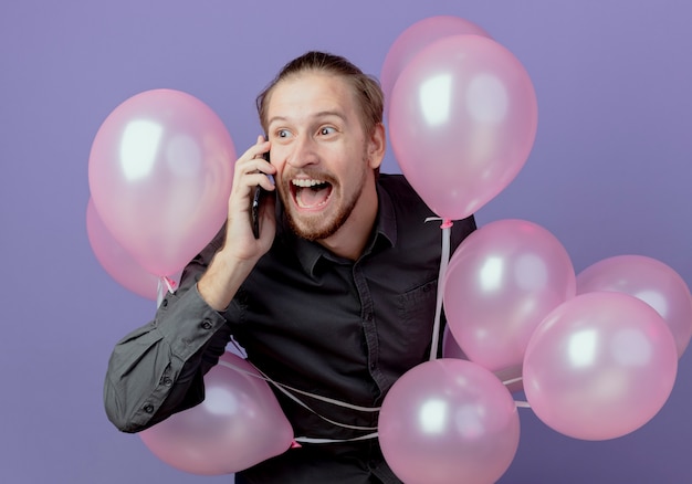 Aufgeregter gutaussehender Mann steht mit Heliumballons, die am Telefon lokalisiert auf lila Wand sprechen
