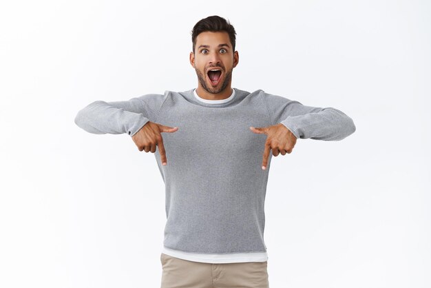 Aufgeregter hübscher junger moderner bärtiger Mann im grauen Pullover mit offenem Mund wunderte sich und amüsierte sich und sagte wow, während die starre Kamera sprachlos vor Nervenkitzel und Faszination die Verwendung des Promo-Codes nach unten empfiehlt