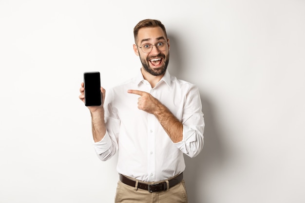 Aufgeregter hübscher Kerl, der Handy zeigt, Finger auf Bildschirm zeigt und lächelt, vor weißem Hintergrund stehend.