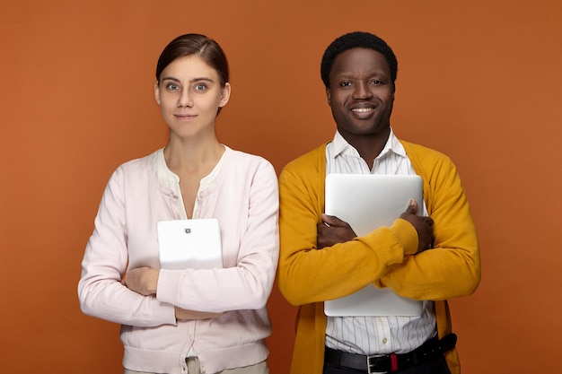 Aufgeregter junger Afroamerikaner männlicher Praktikant und seine attraktive süße Mitarbeiterin posieren nebeneinander mit digitalem Tablet und Laptop, bereit für ihren ersten Arbeitstag, glücklich lächelnd