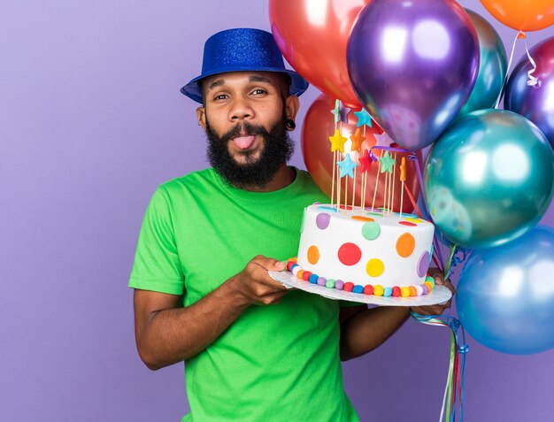 Aufgeregter junger afroamerikanischer Mann mit Partyhut, der Luftballons mit Kuchen hält, der die Zunge isoliert auf blauer Wand zeigt