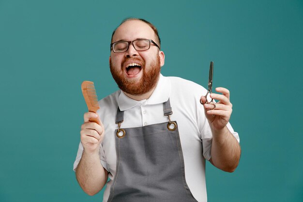 Aufgeregter junger Friseur in Uniform und Brille mit Kamm und Schere mit geschlossenen Augen isoliert auf blauem Hintergrund