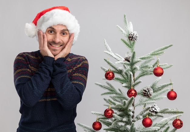 Aufgeregter junger kaukasischer Mann, der Weihnachtshut trägt, der nahe Weihnachtsbaum steht, hält Hände auf Gesicht, das Kamera lokalisiert auf weißem Hintergrund betrachtet