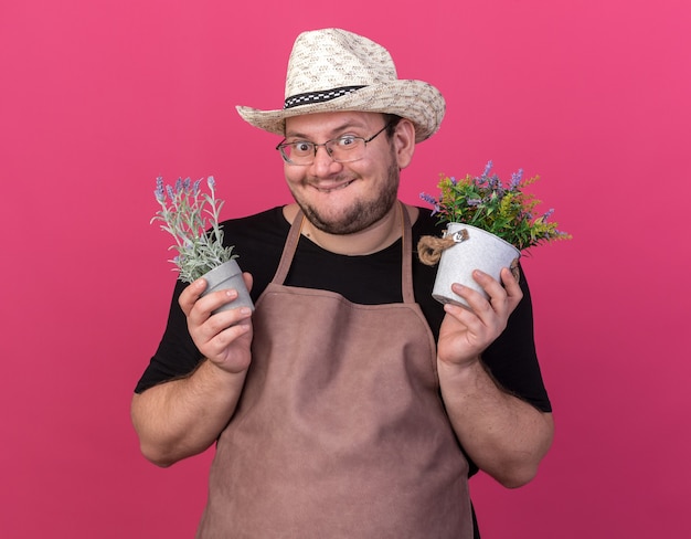 Aufgeregter junger männlicher Gärtner, der Gartenhut trägt, der Blumen in den Blumentöpfen hält, die auf rosa Wand lokalisiert werden