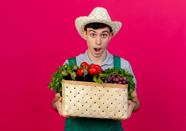 Aufgeregter junger männlicher Gärtner, der Gartenhut trägt, hält Gemüsekorb