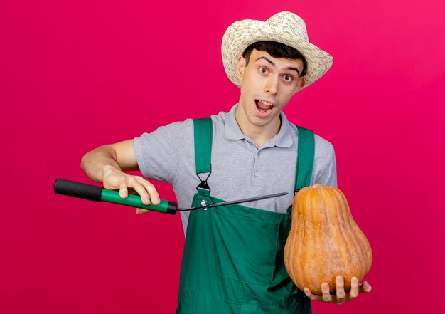 Aufgeregter junger männlicher Gärtner mit Gartenhut hält Klipper und Kürbis