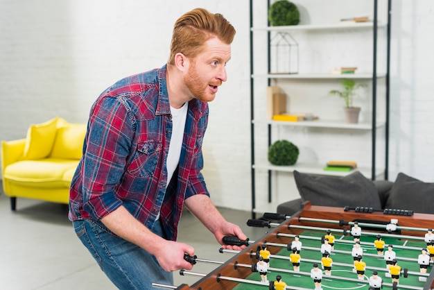 Aufgeregter junger Mann, der das Fußballtischfußballspiel im Wohnzimmer spielt