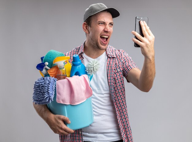 Aufgeregter junger Mann, der eine Kappe trägt, die einen Eimer mit Reinigungswerkzeugen hält, spricht am Telefon isoliert auf weißem Hintergrund
