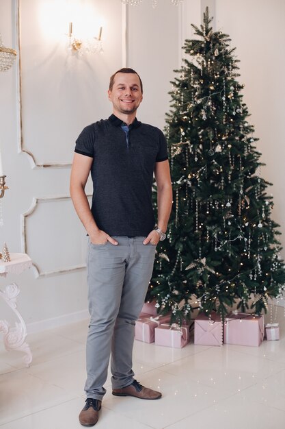 Aufgeregter junger Mann mit den Händen in den Taschen, die festlich fühlen, während sie nahe einem Weihnachtsbaum aufwerfen. Urlaubskonzept