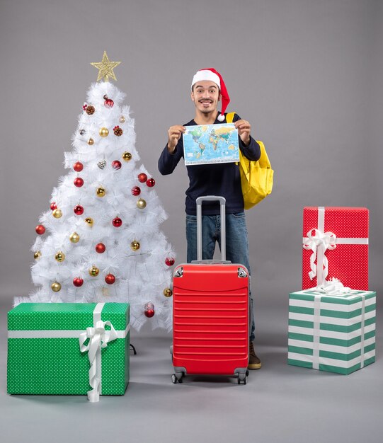 aufgeregter junger Mann mit gelbem Rucksack, der Karte mit beiden Händen nahe weißem Weihnachtsbaum auf grau hält