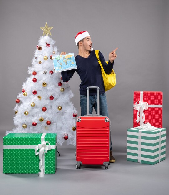 aufgeregter junger Mann mit gelbem Rucksack, der Karte nahe Weihnachtsbaum auf grau hält