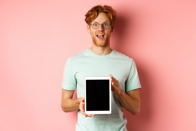 Aufgeregter junger Mann mit roten Haaren und Bart, der Online-Werbung mit digitalem Tablet-Bildschirm auscheckt...