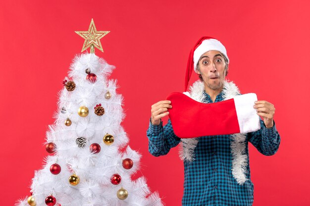 Aufgeregter junger Mann mit Weihnachtsmannhut in einem blauen gestreiften Hemd und Weihnachtssocke nahe Weihnachtsbaum