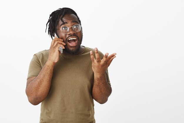 aufgeregter Kerl mit Brille, die mit seinem Telefon gegen die weiße Wand posiert