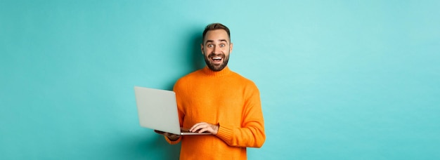 Aufgeregter lächelnder Mann, der am Laptop arbeitet und in die Kamera starrt, glücklich im orangefarbenen Pullover gegen tur