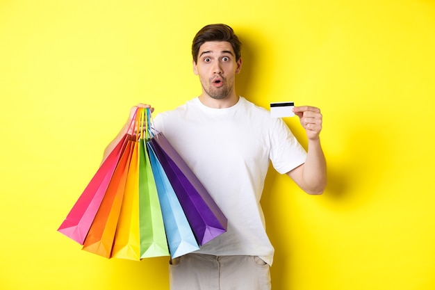 Aufgeregter Mann, der am schwarzen Freitag einkauft, Papiertüten und Kreditkarte hält und vor gelbem Hintergrund steht
