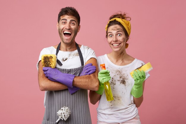 Aufgeregter Mann in Freizeitkleidung, der die Hände gekreuzt hält und einen schmutzigen Schwamm hält, der sich über seine Arbeit freut. Lächelnde Frau, die gelbes Stirnband und weißes T-Shirt hält, das Waschmittel- und Schwammwaschfenster hält