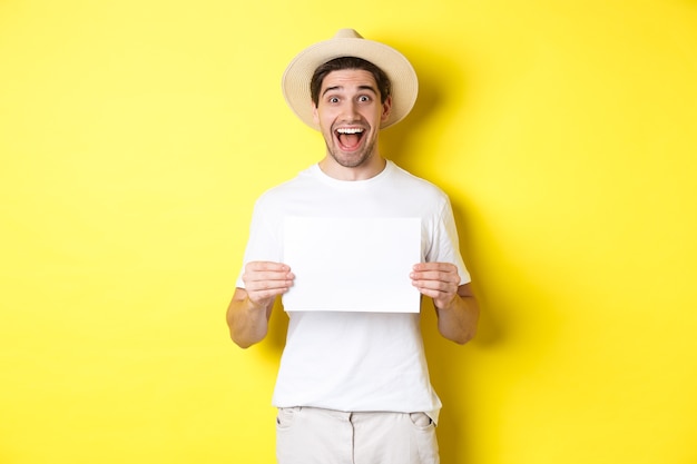 Aufgeregter Tourist, der Ihr Logo oder Zeichen auf leerem Stück Papier zeigt, erstaunt lächelt und vor gelbem Hintergrund steht.