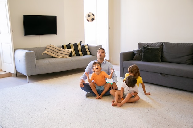 Aufgeregter vater, der mit kindern auf teppich sitzt und spielt. netter verspielter junge, der ball wirft und ihn ansieht. schöne kinder spielen mit papa zu hause. konzept für kindheit, spiel und vaterschaft