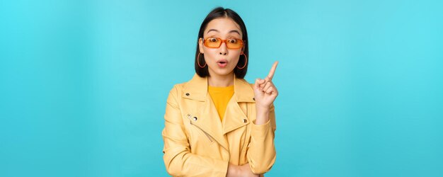 Aufgeregtes asiatisches Mädchen, das den Finger hebt, was darauf hindeutet, dass ein Eureka-Zeichen einen Plan hat, der in einer Sonnenbrille über blauem Hintergrund steht