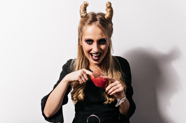 Aufgeregtes blondes Mädchen, das an Halloween Blut trinkt. Atemberaubender Vampir im schwarzen Kleid, das am Karneval aufwirft.